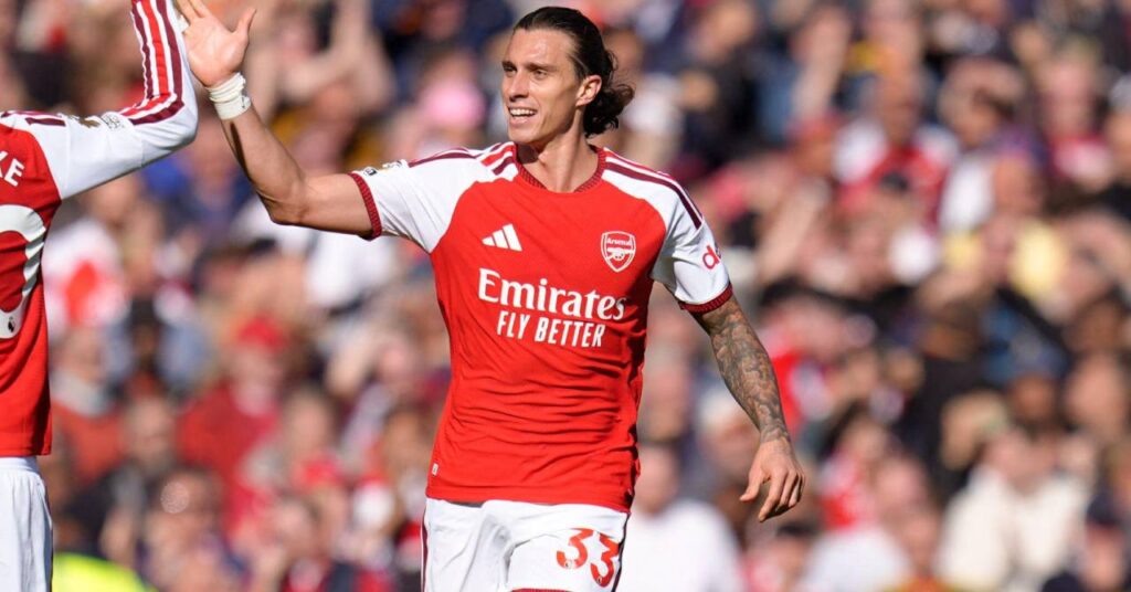 Italy footballer Riccardo Calafiori raising his right arm and smiling to celebrate a goal for Premier League team Arsenal Football Club in a red home shirt with white sleeves and white shorts with red trim and the words Emirates Fly Better, Adidas logo and club crest on the front in white and his number 33 in red on the shorts with the raised right arm of teammate Noni Madueke visible to the left and a blurrily visible crowd in the background.