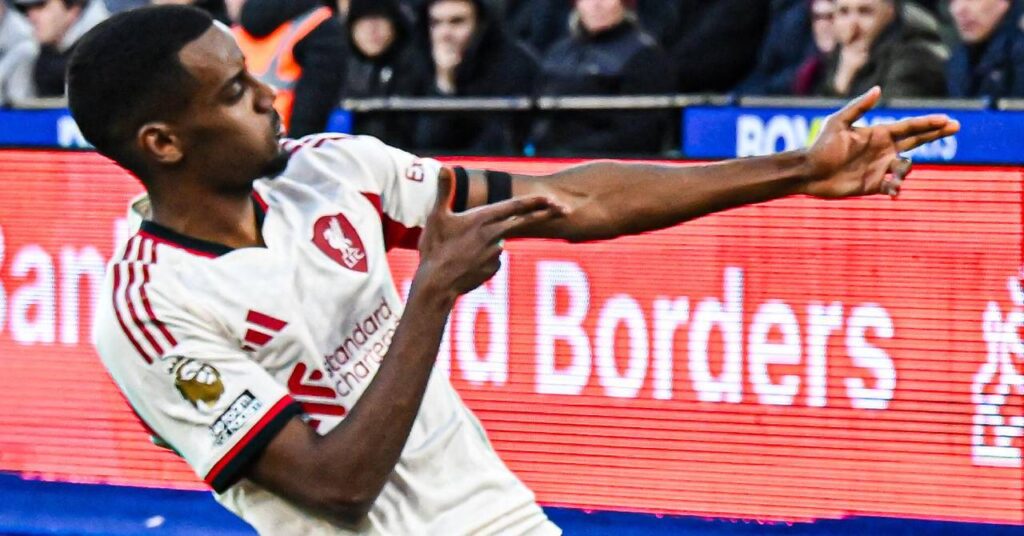 Sweden footballer Alexander Isak kneeling and raising both hands to celebrate a goal while playing for Premier League team Liverpool Football Club in a white kit with red trim in front of a crowd in a stadium